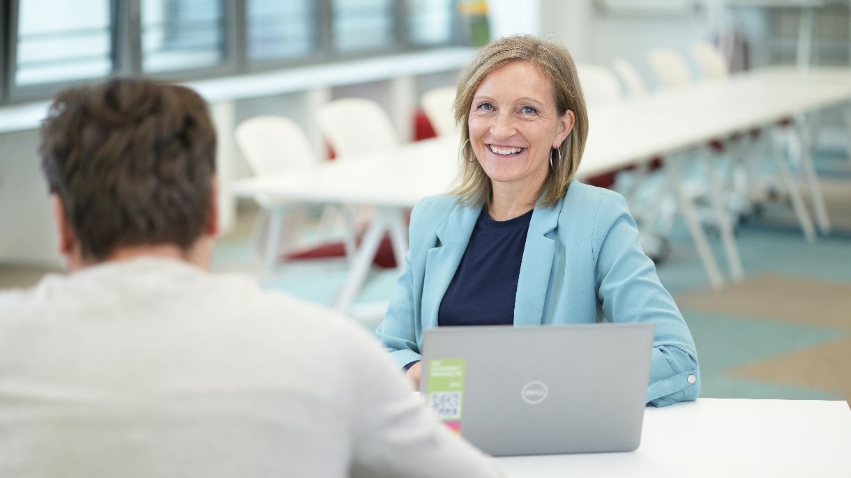 Frau im Anzug sitzt mit Laptop bei Tisch und lächelt in die Kamera.