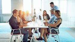 Gruppe von BRZ-Mitarbeiterinnen und Mitarbeitern sitzt an einem Tisch bei der Arbeit in der Innovation Factory. Oranges Licht durchdringt atmosphärisch die lockere, aber produktive Arbeit.