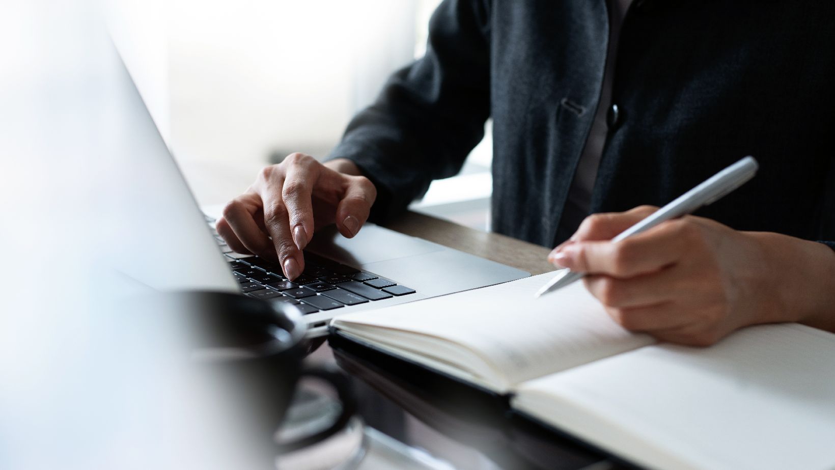 Person arbeitet am Laptop und schreibt in ein Notizbuch