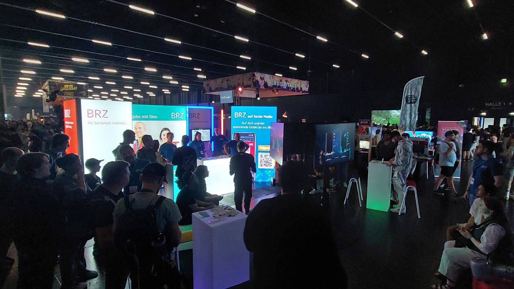 Foto von Standbesucherinnen und Standbesuchern der BRZ-Gaming Area auf der LEVEL UP Messe in Salzburg. Der BRZ-Messestand befindet sich in einer dunklen Veranstaltungshalle, der BRZ-Messestand sticht durch seine LED-Beleuchtung heraus. 